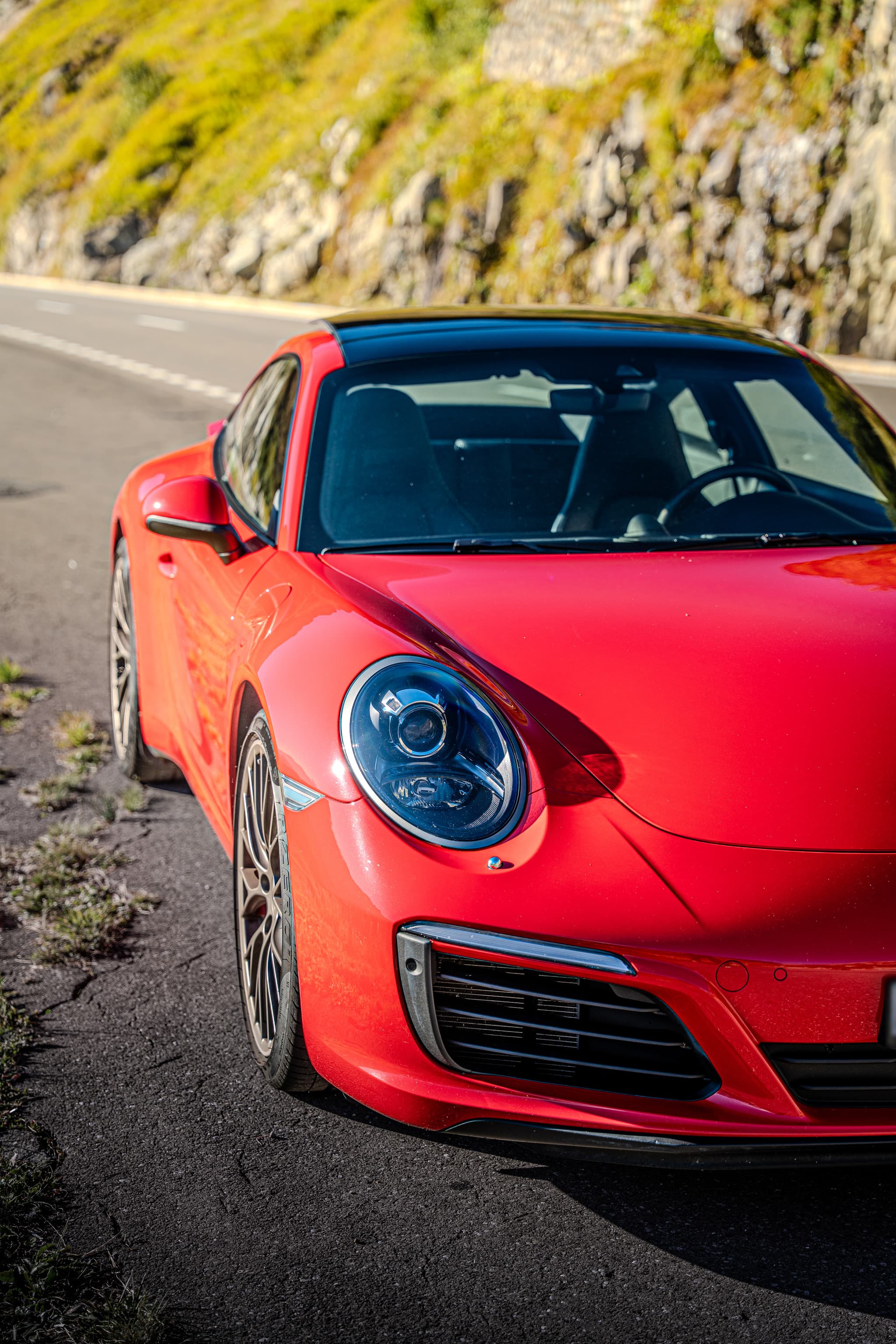 Roter Porsche 911 Carrera S Coupé, Scheinwerfer und Front aus nächster Nähe am Strassenrand, dahinter ein grüner Hang