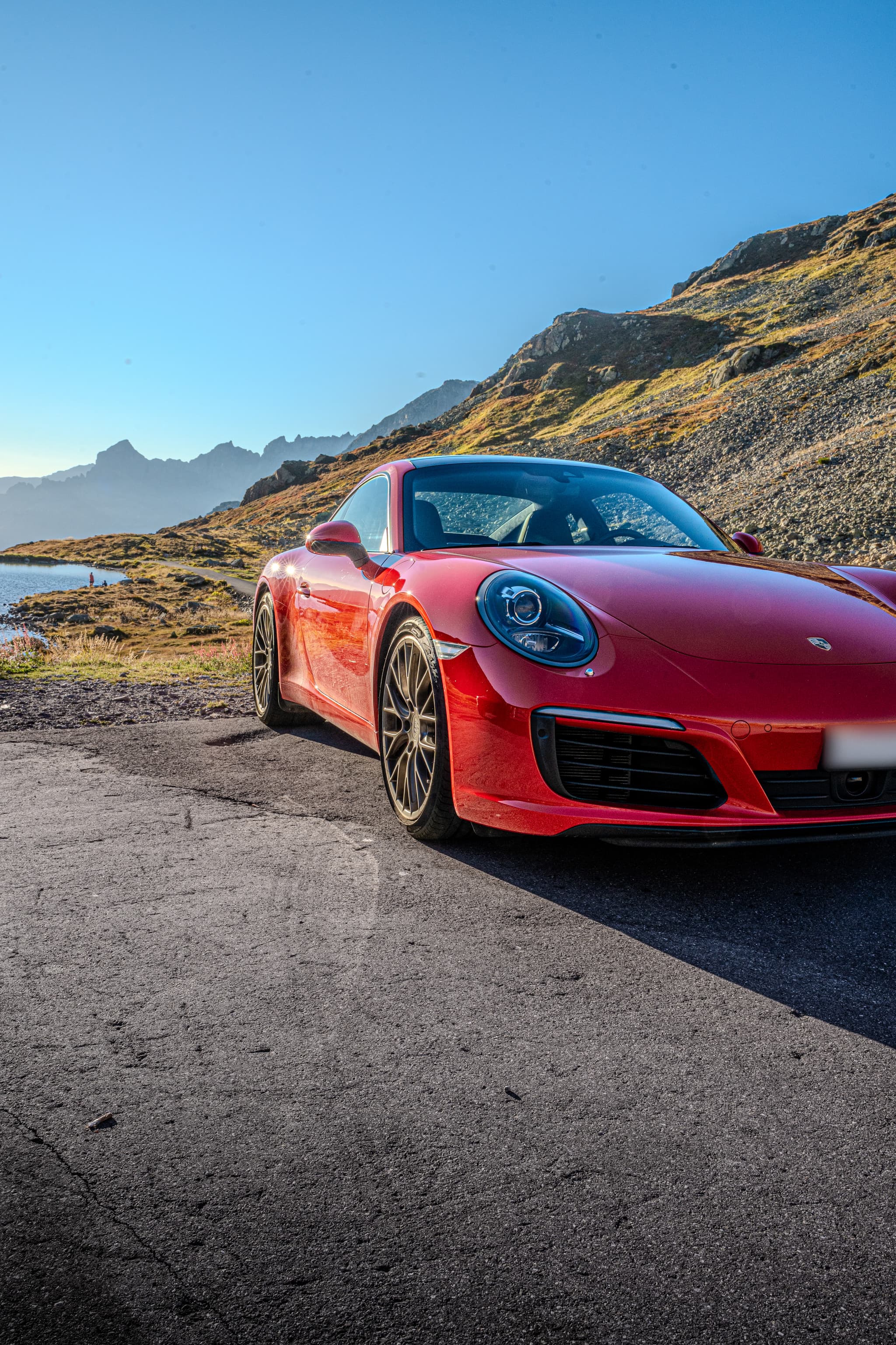 Roter Porsche 911 Carrera S Coupé, Ansicht von schräg vorne aus der Nähe, links ein Bergsee, dahinter ein sonniger Geröllhang