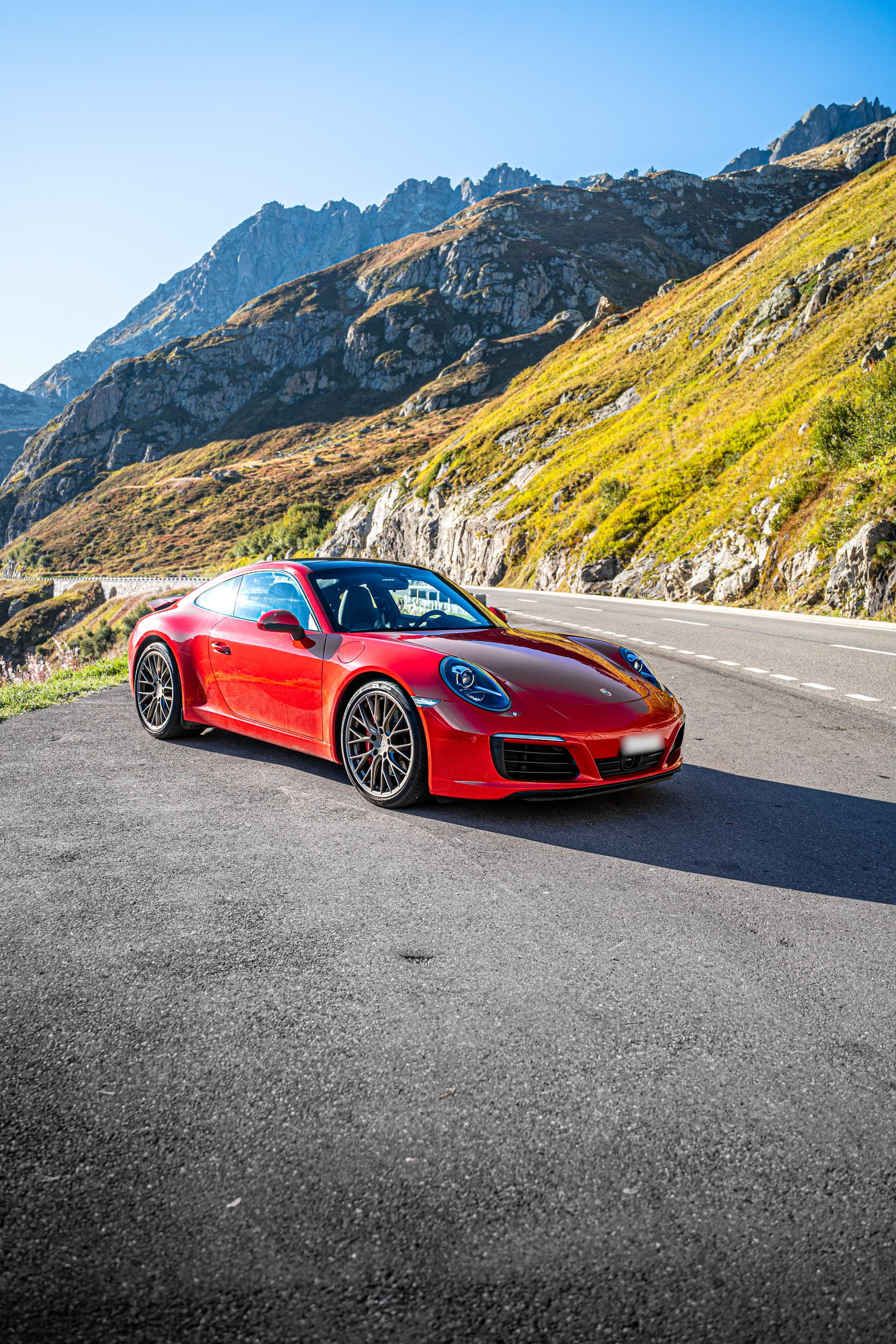 Roter Porsche 911 Carrera S Coupé, Ansicht von schräg vorne auf einer Passstrasse zwischen Felsen und sonnigen Berghängen