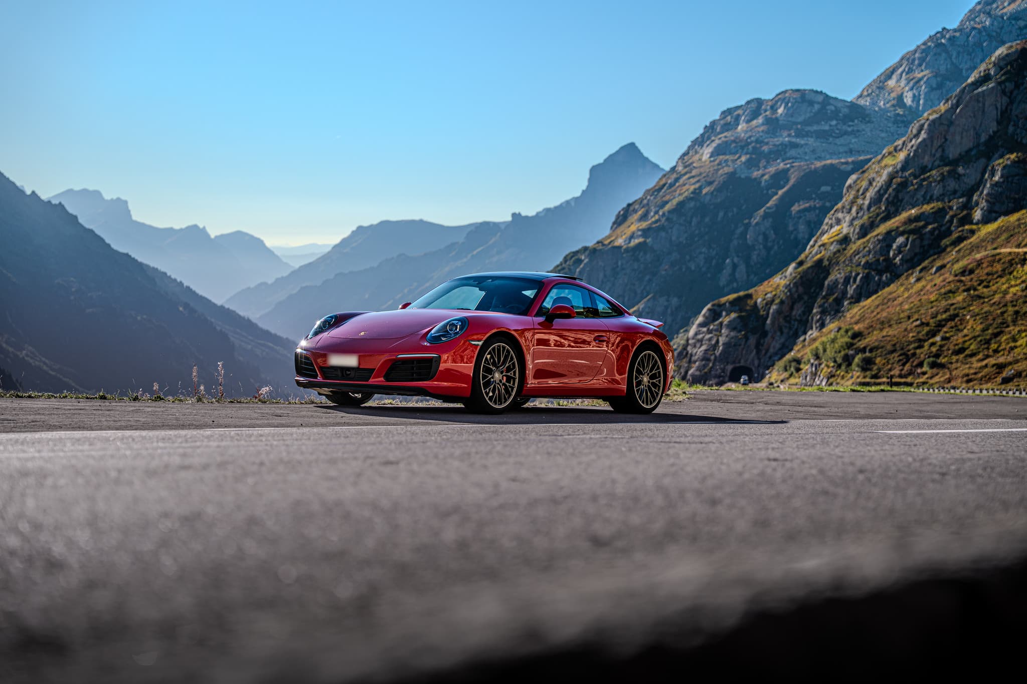 Roter Porsche 911 Carrera S Coupé, Ansicht von schräg vorne auf einer Passstrasse, dahinter gestaffelte Bergketten im Gegenlicht