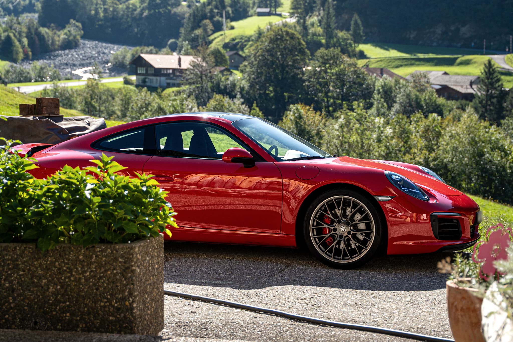 Roter Porsche 911 Carrera S Coupé, Seitenansicht auf einem sonnigen Vorplatz mit Blumenkübeln, dahinter ein grünes Tal mit Chalets