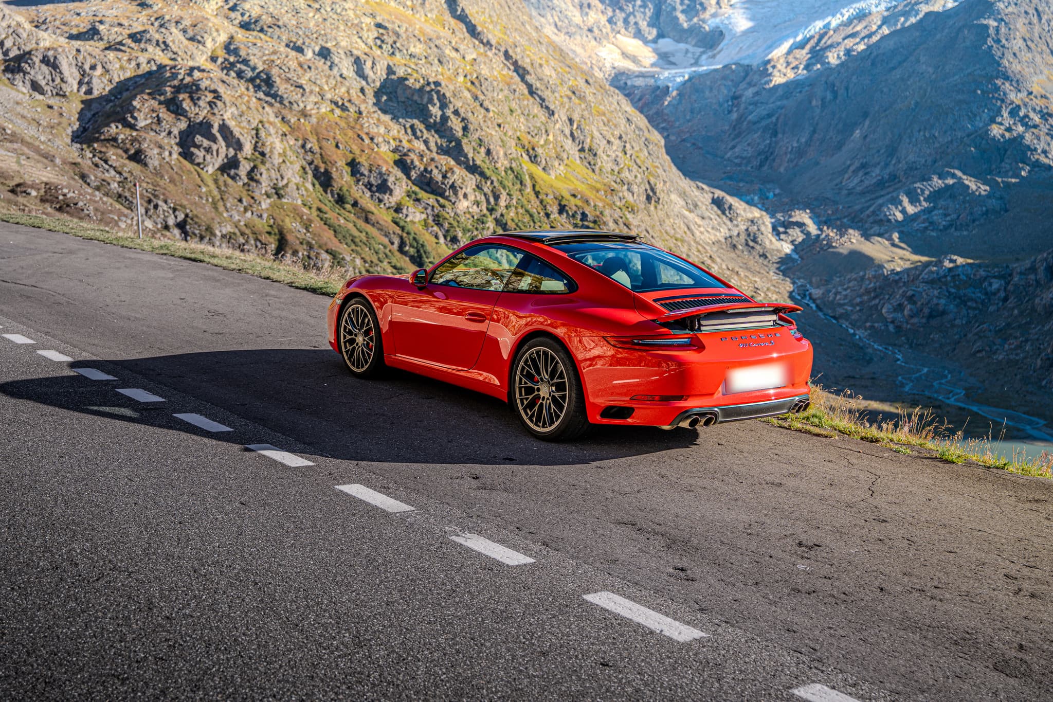 Roter Porsche 911 Carrera S Coupé, Ansicht von schräg hinten auf einer Passstrasse hoch über einem Tal mit Gletscher